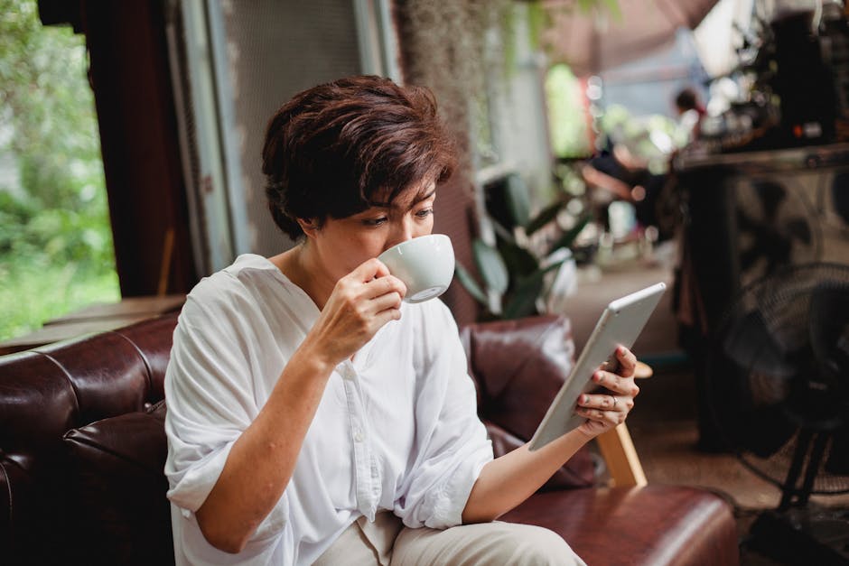 Focused Asian female in trendy clothes drinking cup of beverage and looking at screen of tablet