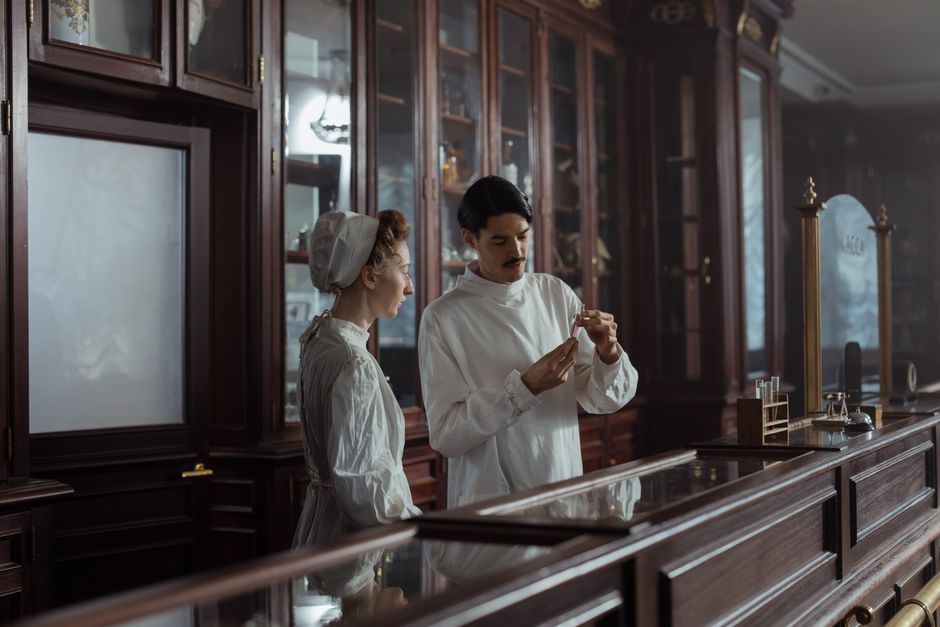 Two pharmacists in vintage attire standing in a classic pharmacy setting, examining medication.