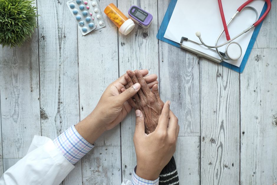 A doctor holding an elderly patient's hands on a desk with medication and stethoscope.