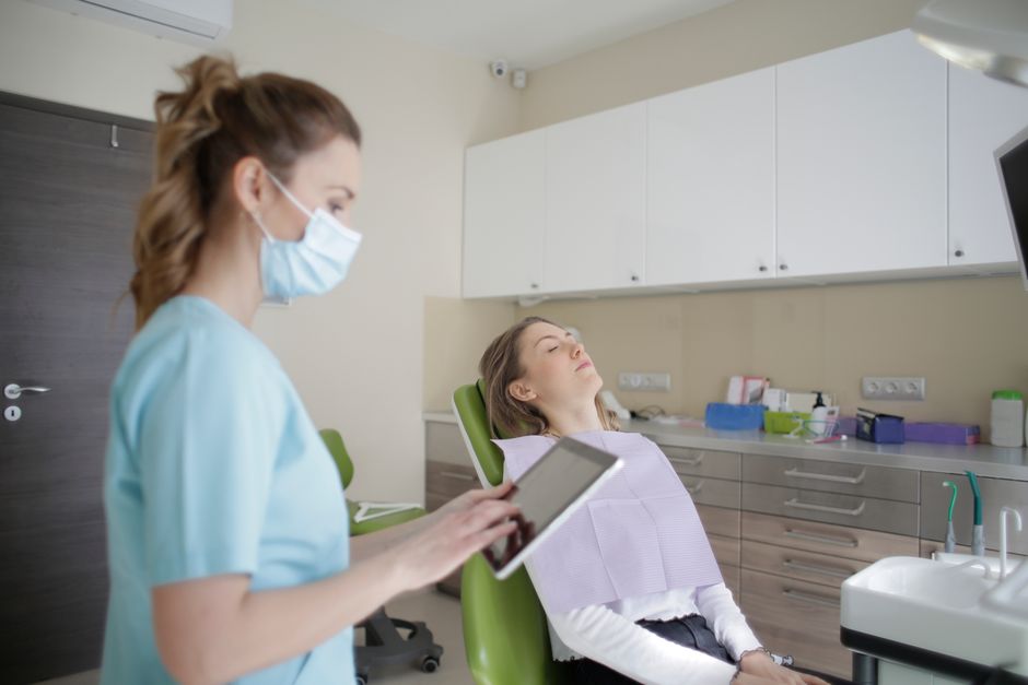 Female dentist in uniform and sterile mask touching tablet screen while standing near female patient sitting on medical chair with closed eyes in afternoon